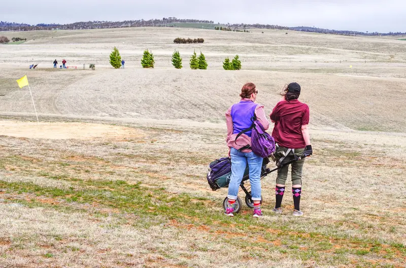 Two women on a golf course carrying bags, enjoying a casual outing in a rural setting