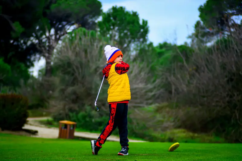 A child golfer in winter clothing swings a club on a lush green golf course